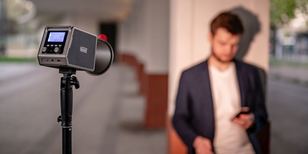 Mężczyzna w granatowej marynarce stoi na zewnątrz budynku, patrząc w dół na telefon w dłoni. Przed nim na czarnym statywie stoi lampa Newell z kwadratowym korpusem, wyświetlaczem pokazującym „5”, dużym czarnym reflektorem i czerwoną osłoną.