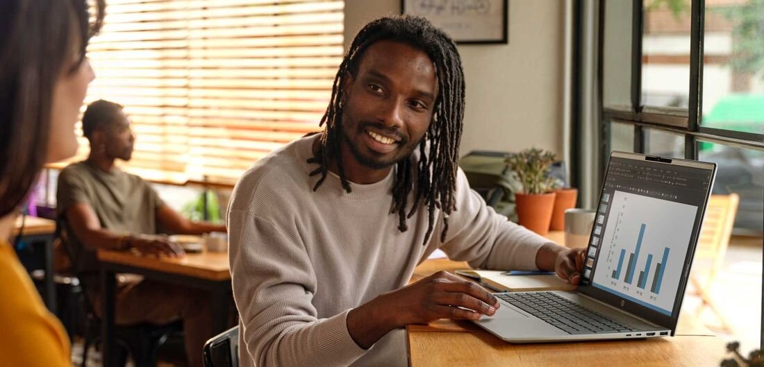 Mężczyzna z dreadami w beżowym swetrze uśmiechający się podczas pracy na laptopie HP Pavilion 16 w kawiarni, na ekranie kolorowe wykresy słupkowe, w tle kobieta przy innym stoliku, ciepła atmosfera pracy zdalnej.