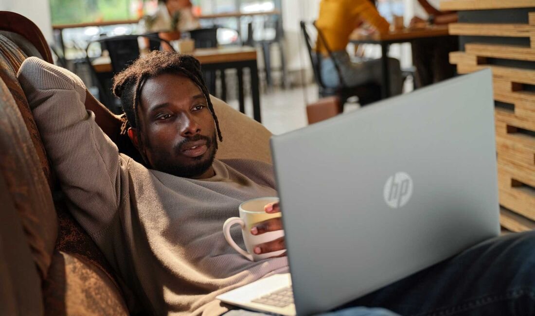 Mężczyzna z dreadami w beżowej bluzie relaksujący się z laptopem HP Pavilion 16 w kawiarni, obok kubek w paski, w tle rozmyte sylwetki innych gości, komfortowe środowisko do pracy i odpoczynku. - Intel Core, HP Fast Charge, 16-calowy ekran