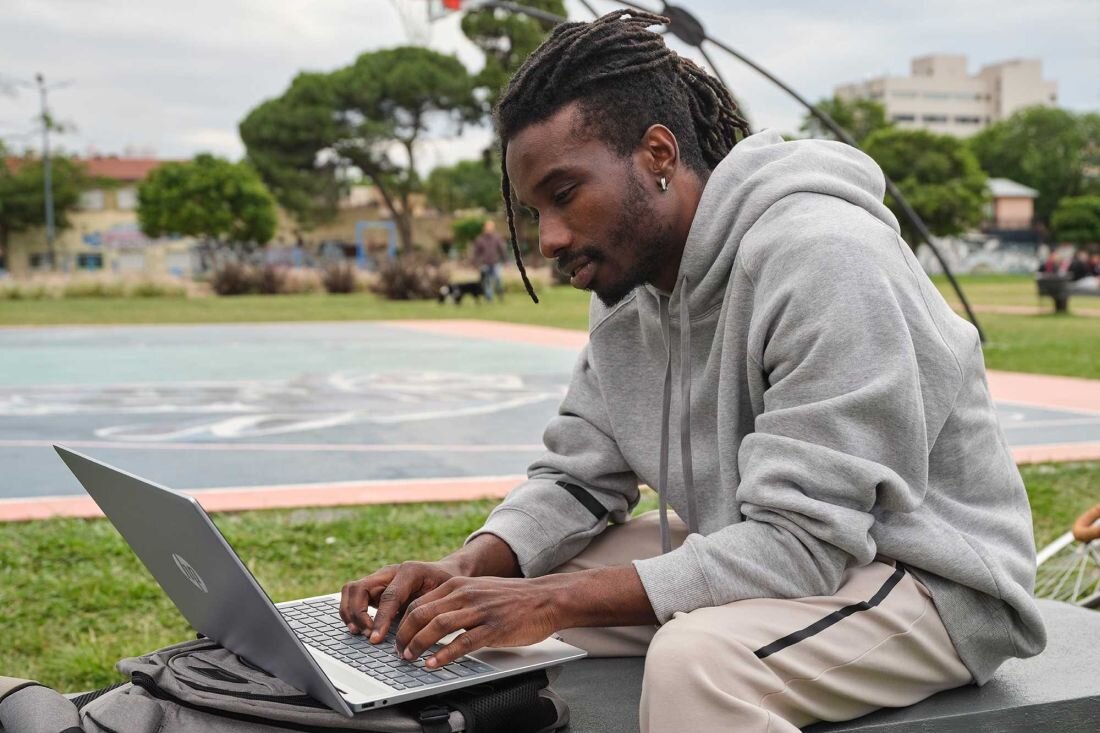 Mężczyzna z dreadami w szarej bluzie z kapturem koncentrujący się podczas pracy na laptopie HP Pavilion 16 w parku miejskim, na ławce obok plecak, w tle kolorowe boisko i zieleń, mobilna praca na świeżym powietrzu. - Intel Core, pamięć LPDDR5, dysk SSD, Intel Iris Xe Graphics