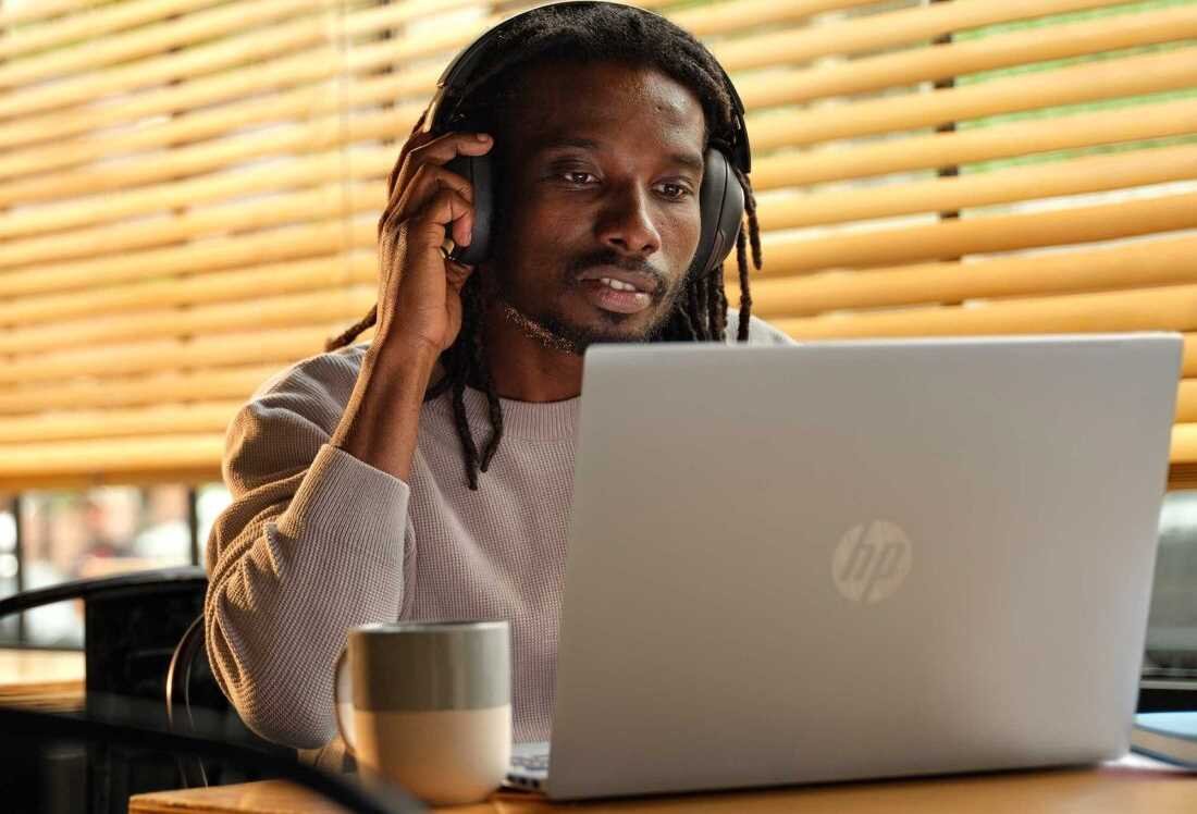 Mężczyzna z dreadami w beżowym swetrze ze słuchawkami na głowie trzymający rękę przy skroni podczas pracy na laptopie HP Pavilion 16 w kawiarni, obok kubek kawy w paski, ciepłe oświetlenie przez żaluzje. - Bateria o pojemności 59 Wh