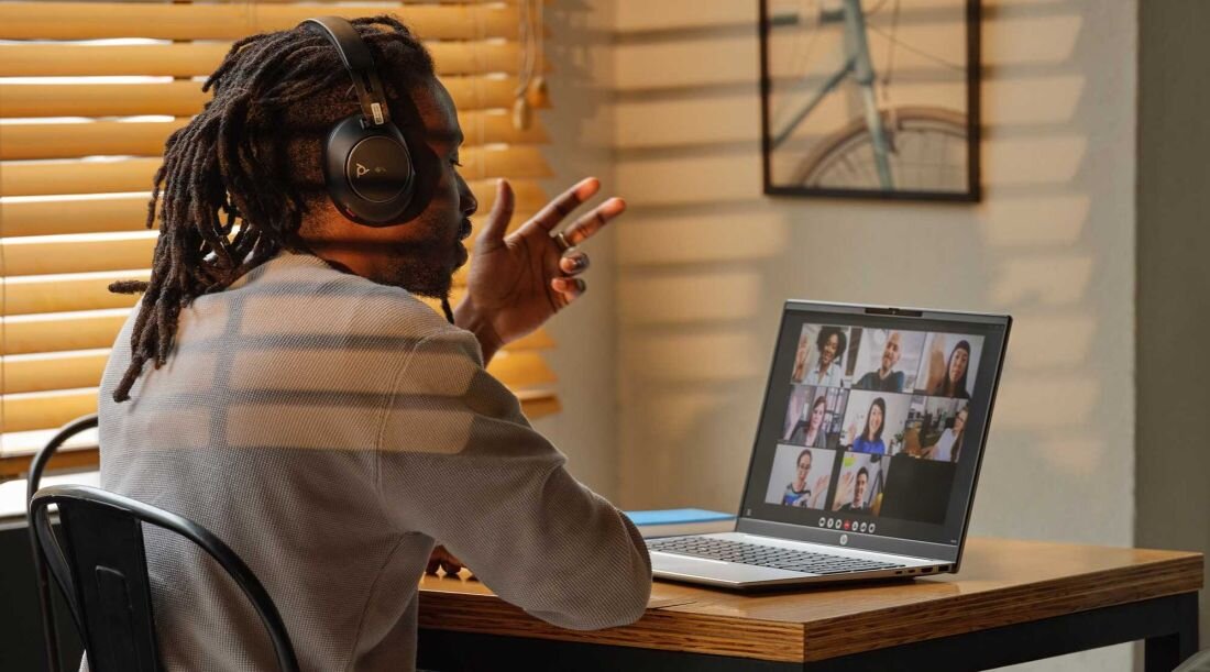 Mężczyzna z dreadami w beżowym swetrze gestykulujący podczas wideokonferencji na laptopie HP Pavilion 16, na ekranie dziewięć uczestników spotkania online w siatce 3x3, w tle żaluzje z ciepłym światłem. - System dwóch głośników HP Audio Boost