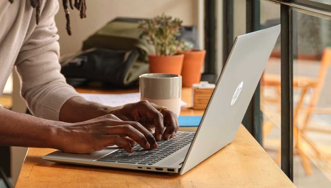 Zbliżenie na dłonie mężczyzny z dreadami pisującego na klawiaturze laptopa HP Pavilion 16, obok kubek kawy w paski i roślina doniczkowa w pomarańczowej doniczce, na drewnianym stole w kawiarni. - Copilot w systemie Windows