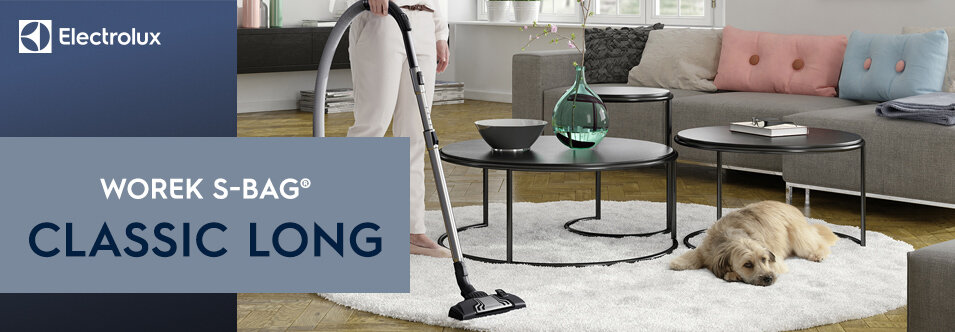 Na obrazie widoczny jest odkurzacz na białym dywanie w salonie z psem leżącym obok. Po lewej stronie znajduje się niebieski panel z logo Electrolux i tekstem: \'WOREK S-BAG® CLASSIC LONG\'.