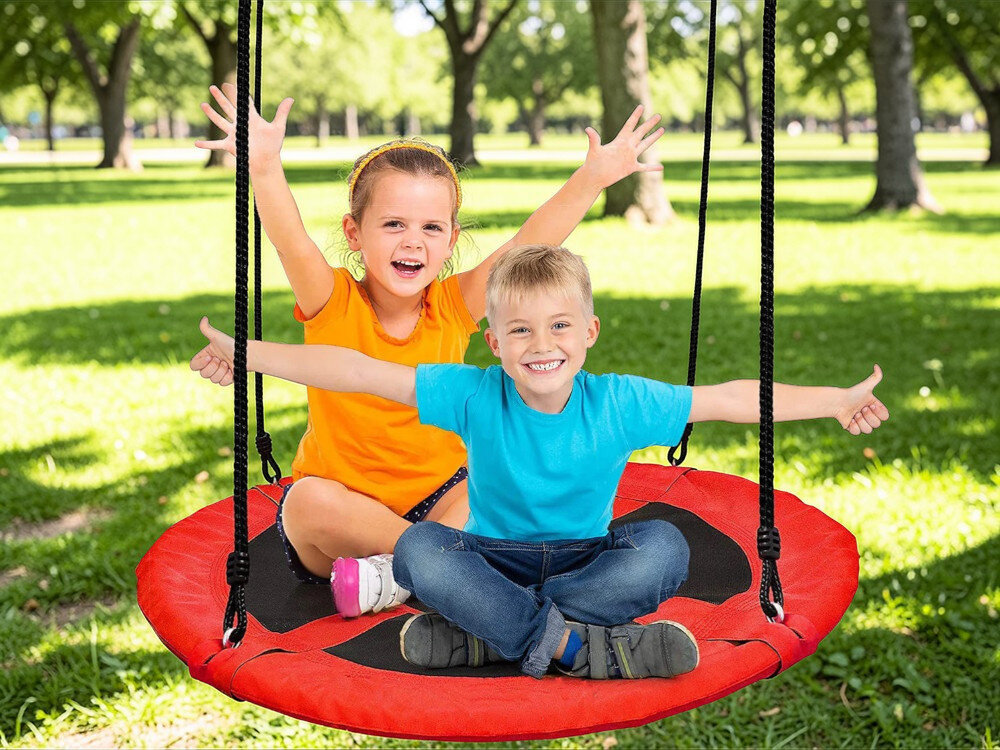 Dwoje dzieci siedzi na czerwonej huśtawce ogrodowej w kształcie bocianiego gniazda, otoczonej zielonymi drzewami. Dzieci są uśmiechnięte i mają uniesione ręce.
