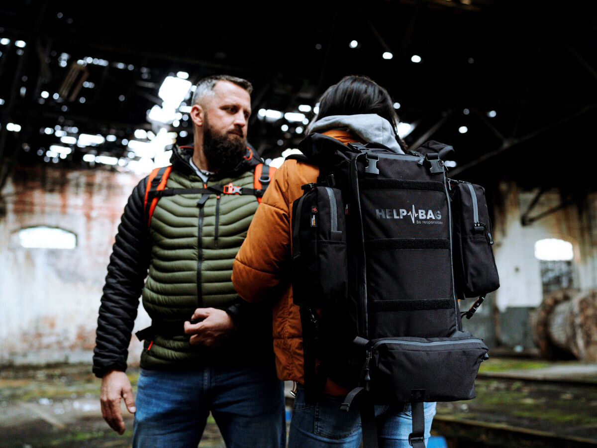 Plecak ewakuacyjny HELP BAG Max Czarny, zagrożenie, ewakuacja, survival, plecak, w tle opuszczona fabryka, kobieta z mężczyzną z plecakami helpbag stoją w środku fabryki
