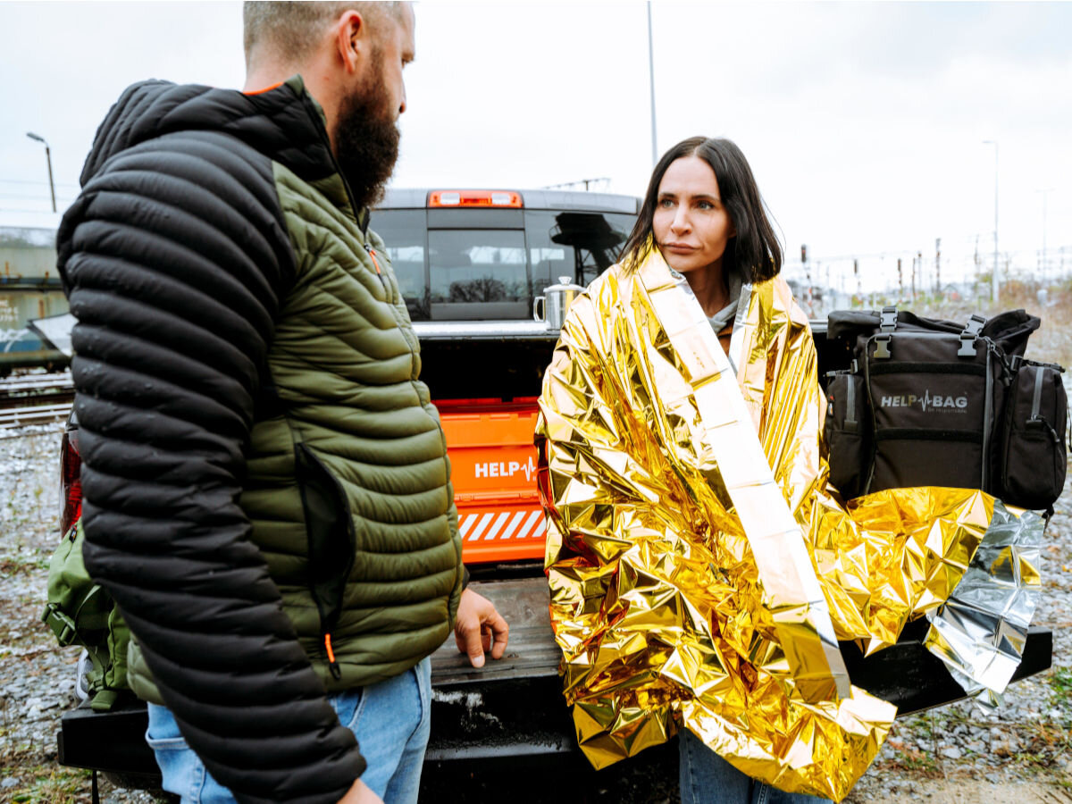 Plecak ewakuacyjny HELP BAG Max Czarny, zestaw, awaria, plecak, kryzys, blackout, wytrzymałość, w tle auto i bagażnik otwarty wraz z skrzynią, kobieta owinięta w folię NRC, obok stoi mężczyzna