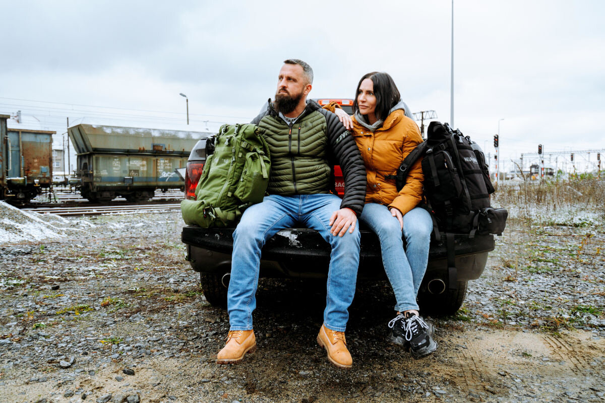 Plecak ewakuacyjny HELP BAG Max Zielony, zestaw, awaria, plecak, kryzys, blackout, wytrzymałość, w tle kolej przemysłowa, para siedzi na bagażniku auta, obok plecak