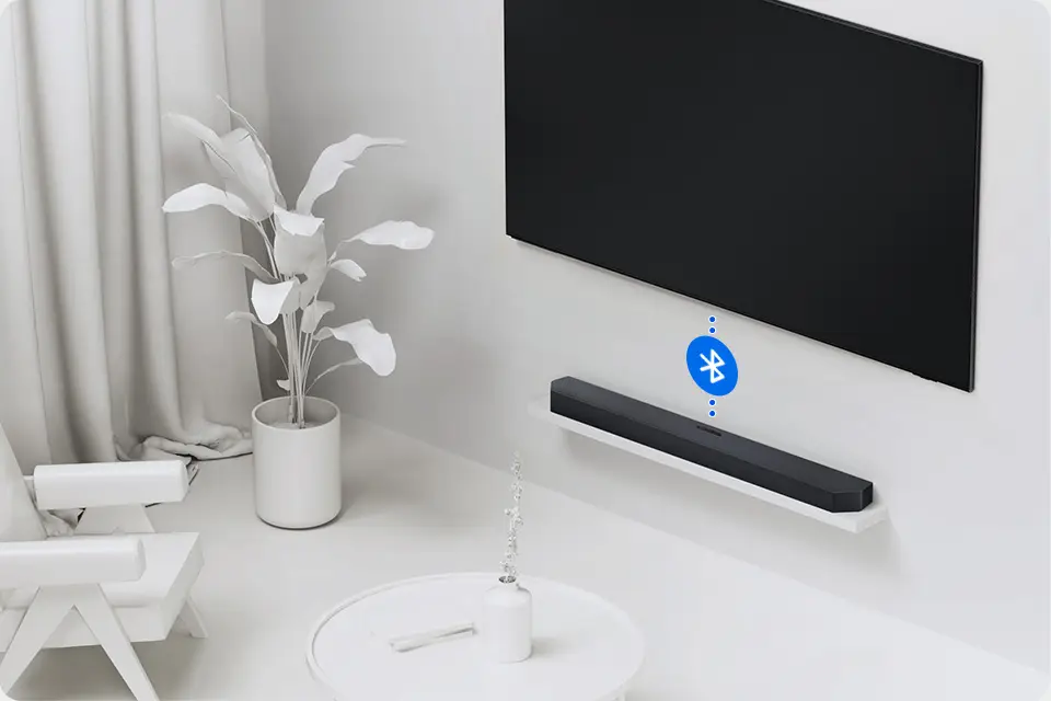 Na ścianie wisi czarny telewizor, poniżej znajduje się czarny soundbar na białej półce. Obok widoczny jest niebieski symbol Bluetooth, a w pomieszczeniu znajduje się biała roślina w doniczce i biały fotel.