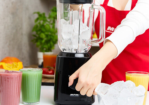 Kobieta w czerwonym fartuchu włącza blender kielichowy MBL-37 wypełniony kostkami lodu. Obok szklanki z sokiem.