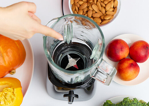 Widok z góry na szklany kielich blendera MBL-37 z ostrzami, wskazywany palcem, otoczony świeżymi składnikami.