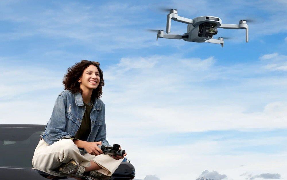 Dron POTENSIC Atom 2 Expansion Kit Uśmiechnięta kobieta siedzi na masce samochodu i trzyma kontroler sterując dronem na tle nieba