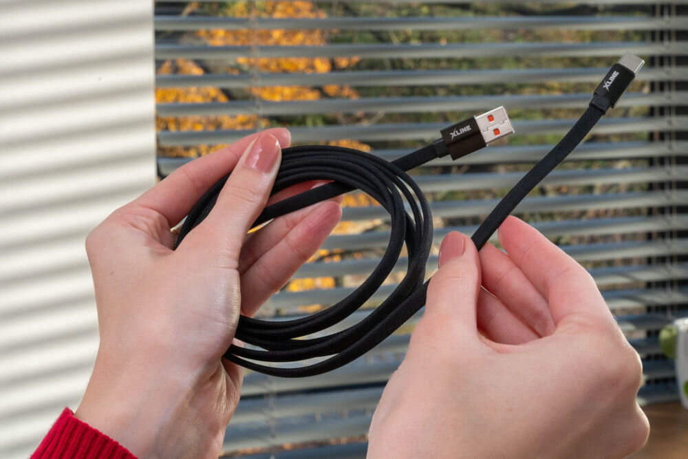 Kabel XLINE UC02KFC-AC USB-A USB-C szybkie ładowanie 60W 2m nylonowy oplot Dłonie trzymające zwinięty czarny kabel USB-C XLINE w wytrzymałym nylonowym oplocie, prezentujące wzmocnione wtyki USB-A oraz USB-C na tle okna.