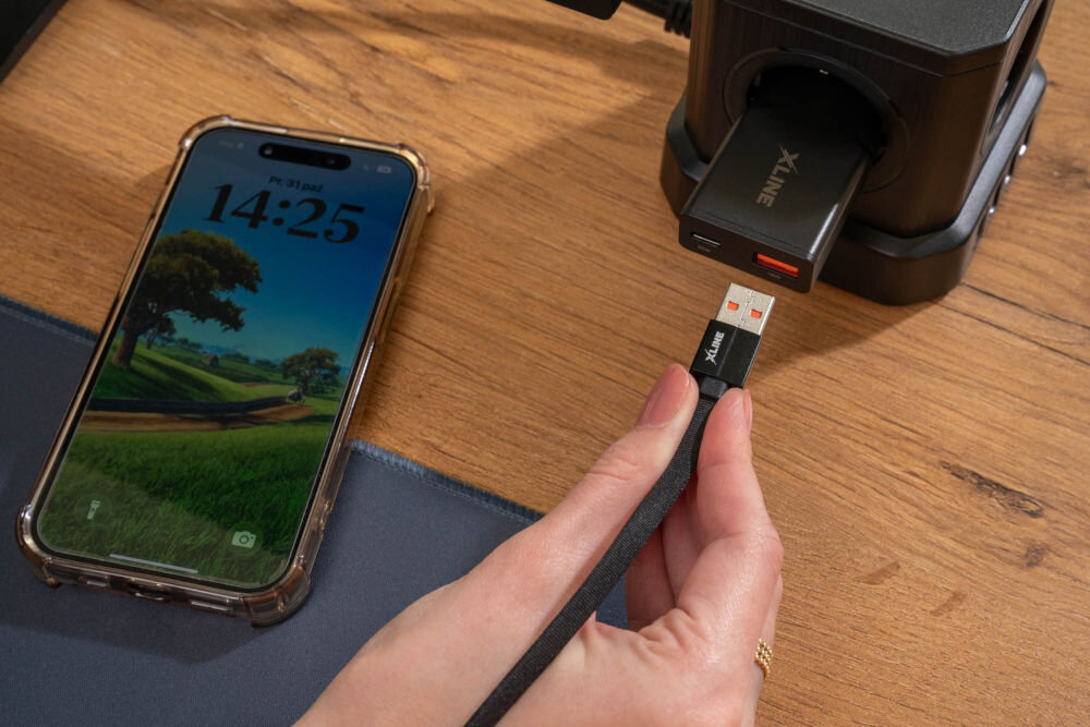 Kabel XLINE UC02KFC-AC USB-A USB-C szybkie ładowanie 60W 2m nylonowy oplot Podłączanie czarnego kabla XLINE do kostki ładowarki sieciowej w gniazdku, obok leżącego smartfona na drewnianym blacie biurka.