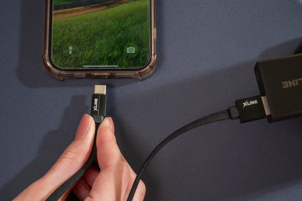 Kabel XLINE UC02KFC-AC nylonowy oplot płaski przewód trwała konstrukcja Zbliżenie z góry na dłoń podłączającą płaski kabel USB-C do portu ładowania smartfona, drugi koniec kabla wpięty do czarnego zasilacza Power Delivery.