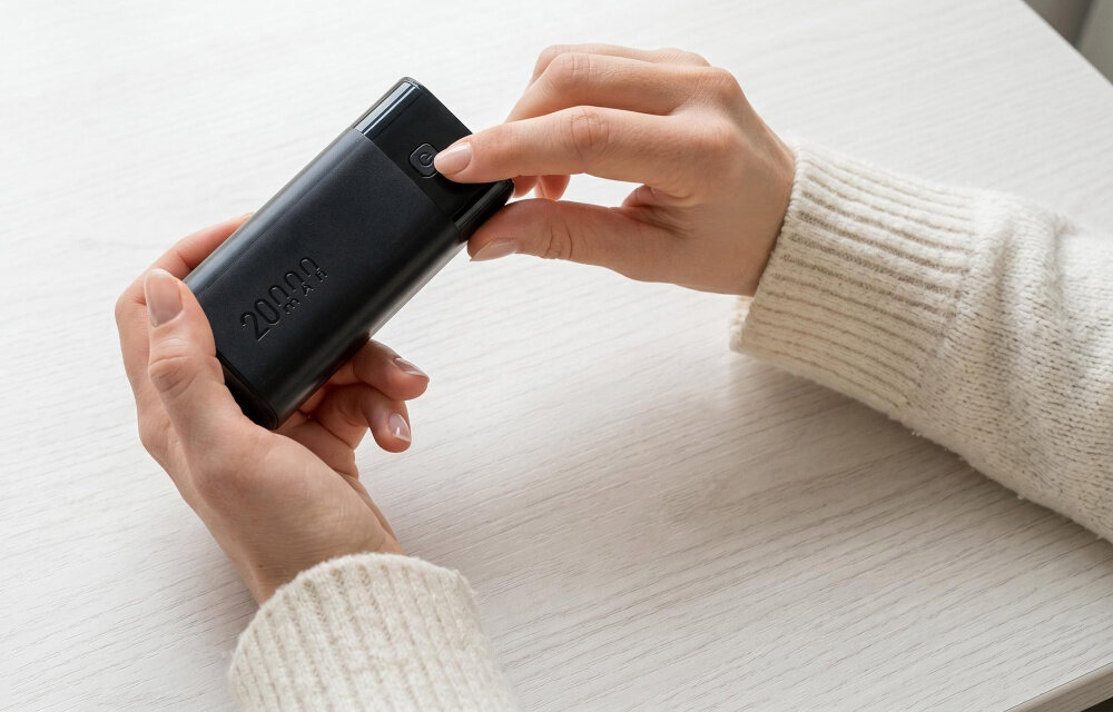 Osoba trzyma czarny powerbank z widocznym napisem \'20000\' na białym stole. Powerbank ma prostokątny kształt i zaokrąglone krawędzie.