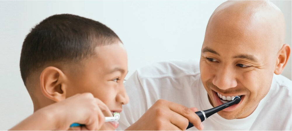 Na zdjęciu widać dwie osoby używające elektrycznych szczoteczek do zębów. Osoby są uśmiechnięte i szczotkują zęby.