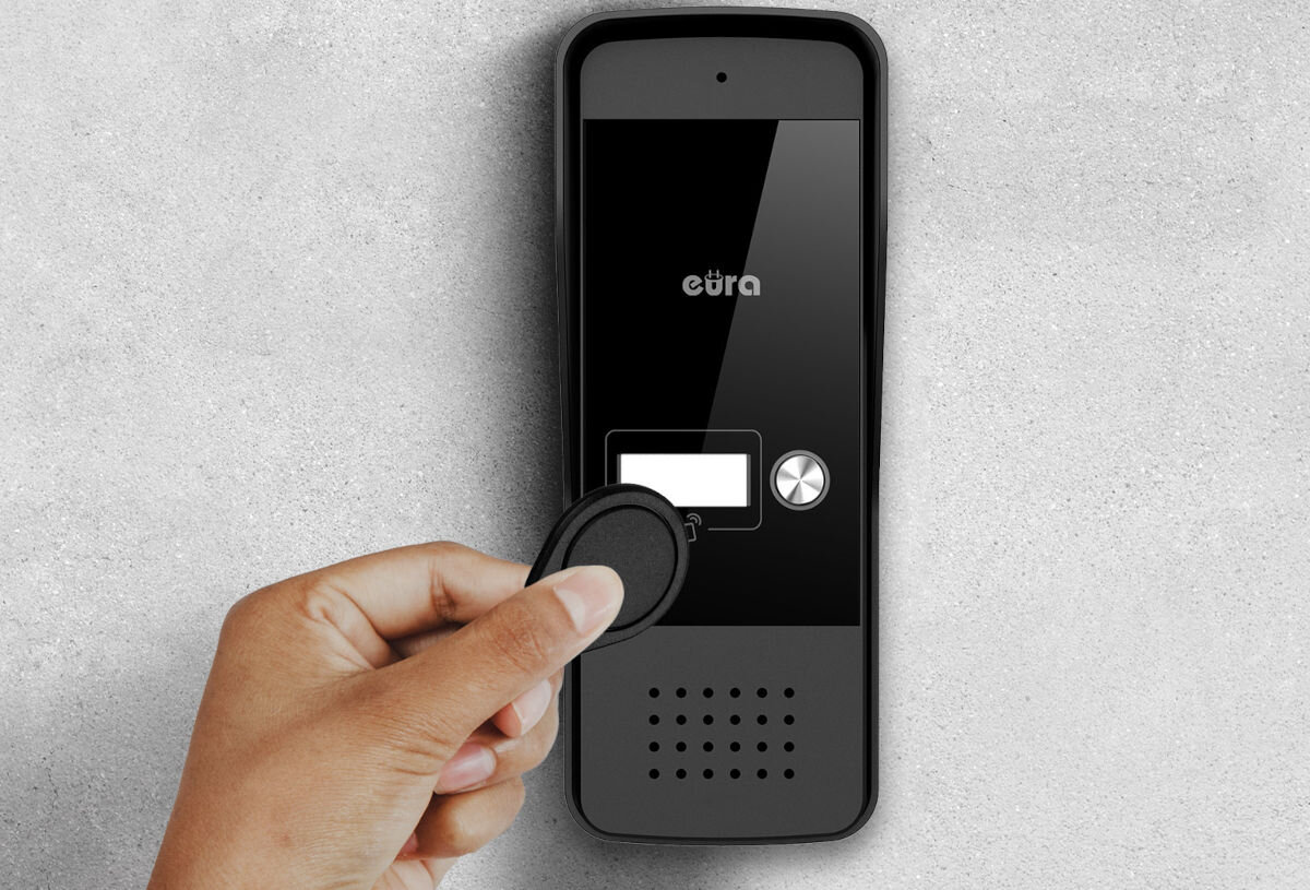 Domofon EURA ADP-25A3 Czarny, RFID, bezdotykowość, brelok, wygoda, Zewnętrzna kaseta domofonowa Eura z panelem dotykowym/czytnikiem RFID, trzymany nad nią czarny brelok zbliżeniowy, na tle jasnej ściany.