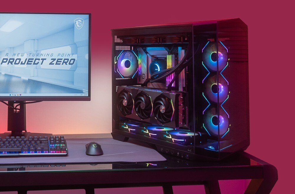Komputer MAD DOG Powered by MSI PANO100-A01DR32 Widok na stanowisko komputerowe z monitorem, myszką i klawiaturą z podświetleniem RGB. Po prawej stronie znajduje się obudowa komputera z przezroczystym bokiem i przodem oraz podświetlanymi wentylatorami, przeznaczenie, cechy, zalety