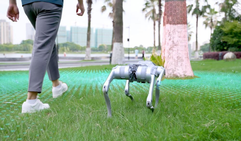 Robot pies UNITREE Go2 Air Szary robot Unitree GO2 idzie po trawie w parku. Część trawy jest wizualizowana jako zielona siatka 3D, a drzewo ma czerwoną siatkę 3D, co sugeruje tryb mapowania i percepcji otoczenia przez robota. Na pierwszym planie widoczne są nogi idącego człowieka.