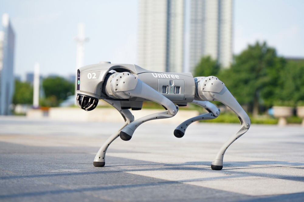 Robot pies UNITREE Go2 Pro Robot Unitree GO2 stoi na brukowanym placu przed nowoczesnymi, wysokimi budynkami. Jest to portret robota na tle miejskiego krajobrazu.
