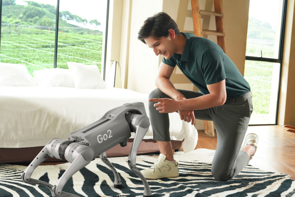 Robot pies UNITREE Go2 Pro Młody mężczyzna, uśmiechnięty i ubrany w zieloną koszulkę polo, klęczy na dywanie, wchodząc w interakcję z czworonożnym robotem Unitree GO2, który stoi przed nim. Scena dzieje się w jasnym, nowoczesnym wnętrzu.