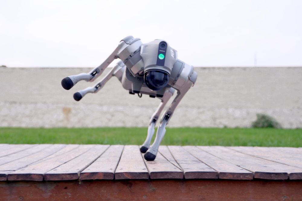 Robot pies UNITREE Go2 Pro Czworonożny, szary robot Unitree GO2 skacze w powietrzu, lądując na drewnianej platformie. Robot jest sfotografowany w trakcie dynamicznego ruchu, z nogami uniesionymi nad platformą