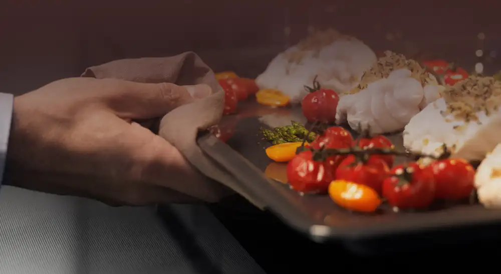 Osoba trzyma blachę z pieczonymi pomidorami koktajlowymi, białymi potrawami i ziołami. Blacha jest wyciągana z piekarnika przy użyciu ściereczki.