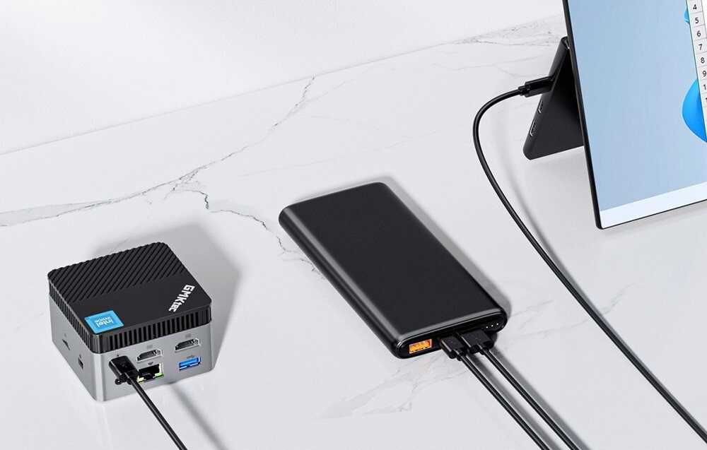Komputer GMKTEC G5 Mały szary komputer podłączony do monitora i czarnego powerbanku leżącego na białym biurku, system, obsługa, zasilanie