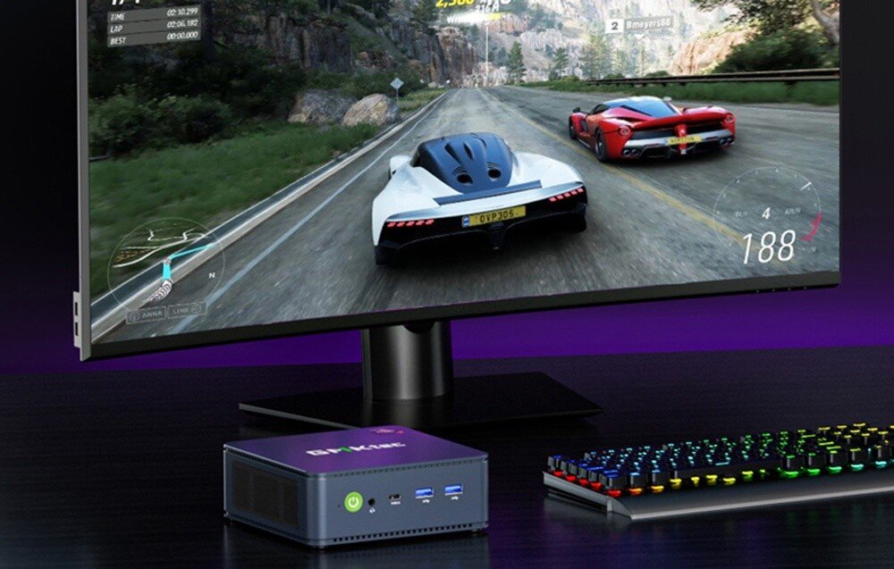Komputer GMKTEC NucBox M5 Plus Mały komputer w ciemnej obudowie stojący na biurku obok monitora wyświetlającego grę wyścigową i klawiatury z podświetleniem RGB, opis