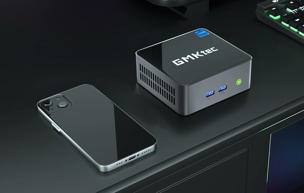 Komputer GMKTEC NucBox M3 Plus Mały komputer w czarnej obudowie stojący na biurku obok smartfona i klawiatury, łączność Wi-Fi, Bluetooth
