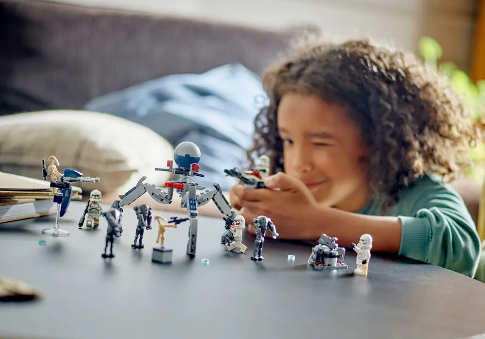 Na stole znajdują się figurki i modele z zestawu LEGO, przedstawiające postacie i maszyny z serii Star Wars. W tle widoczna jest osoba bawiąca się jednym z modeli.