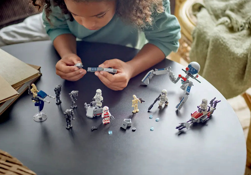 Dziecko bawi się zestawem klocków LEGO przedstawiającym postacie i pojazdy z serii Star Wars na okrągłym stole. Widać różne figurki, w tym żołnierzy i droidy, oraz małe elementy klocków.