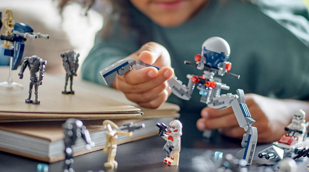 Osoba bawi się zestawem klocków LEGO przedstawiającym postacie i pojazdy z serii Star Wars. Na stole znajdują się różne figurki, w tym droidy i żołnierze.