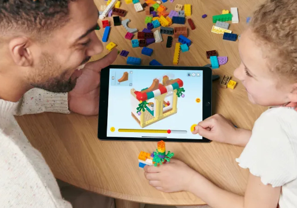 Na stole leży tablet z wyświetlonym modelem konstrukcji z klocków, wokół którego rozrzucone są kolorowe klocki LEGO. Dwie osoby siedzą przy stole, jedna z nich trzyma w ręku zbudowany element z klocków.