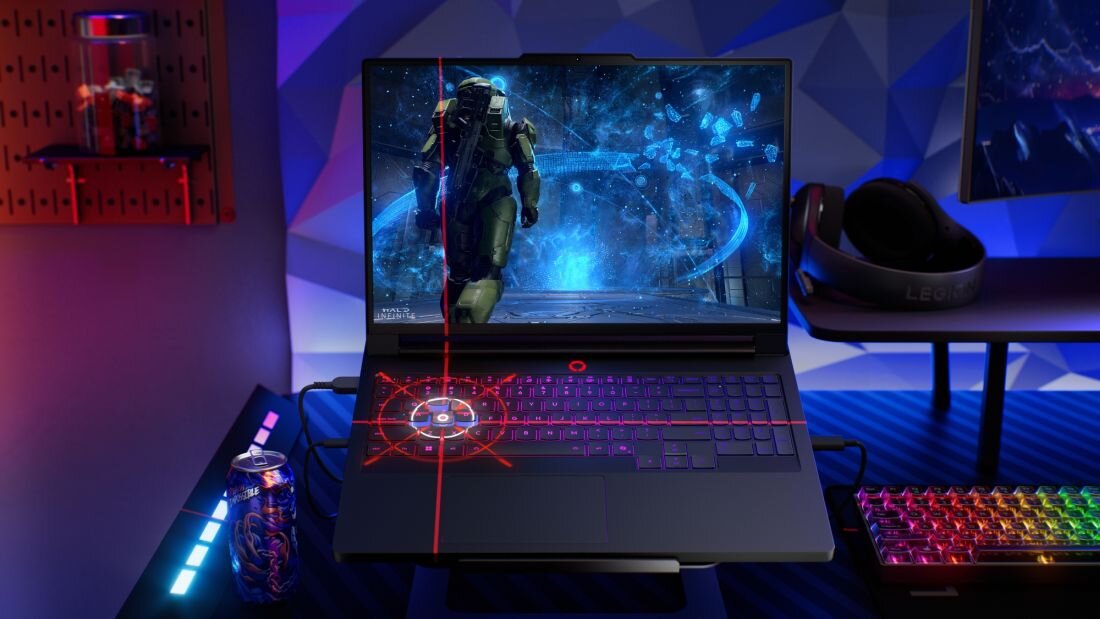 Na biurku znajduje się laptop z wyświetlonym na ekranie obrazem z gry, obok klawiatura z podświetleniem RGB i słuchawki. W tle widoczna jest puszka napoju oraz elementy dekoracyjne.