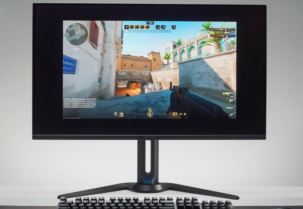 Monitor GIGABYTE Aorus FO27Q2 Monitor na biurku wyświetlający grę FPS. W tle jasne pomieszczenie. Na biurku znajduje się klawiatura mechaniczna, funkcje, przełącznik