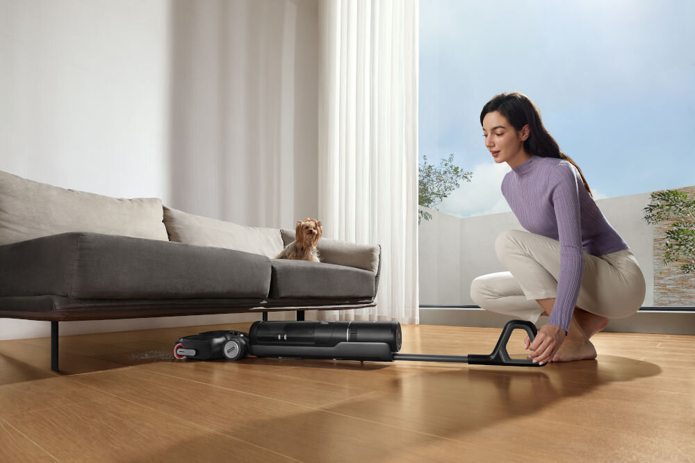 Osoba używa odkurzacza o nowoczesnym designie na drewnianej podłodze w jasnym pomieszczeniu. Na sofie siedzi mały pies, a w tle widać duże okno z zasłonami.