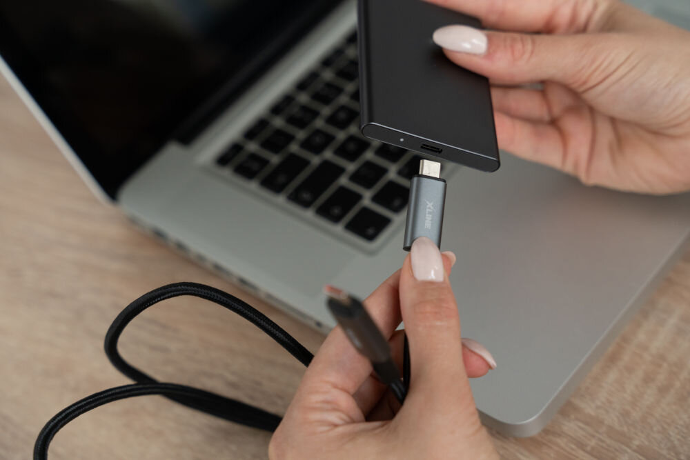 Osoba trzyma czarny kabel USB-C, który jest podłączany do czarnego urządzenia. W tle widoczna jest klawiatura laptopa.