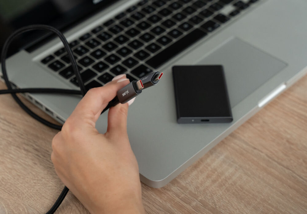 Osoba trzyma czarny kabel USB-C z widocznym logo \'XLINE\' nad klawiaturą laptopa. Obok laptopa leży czarne urządzenie o prostokątnym kształcie.