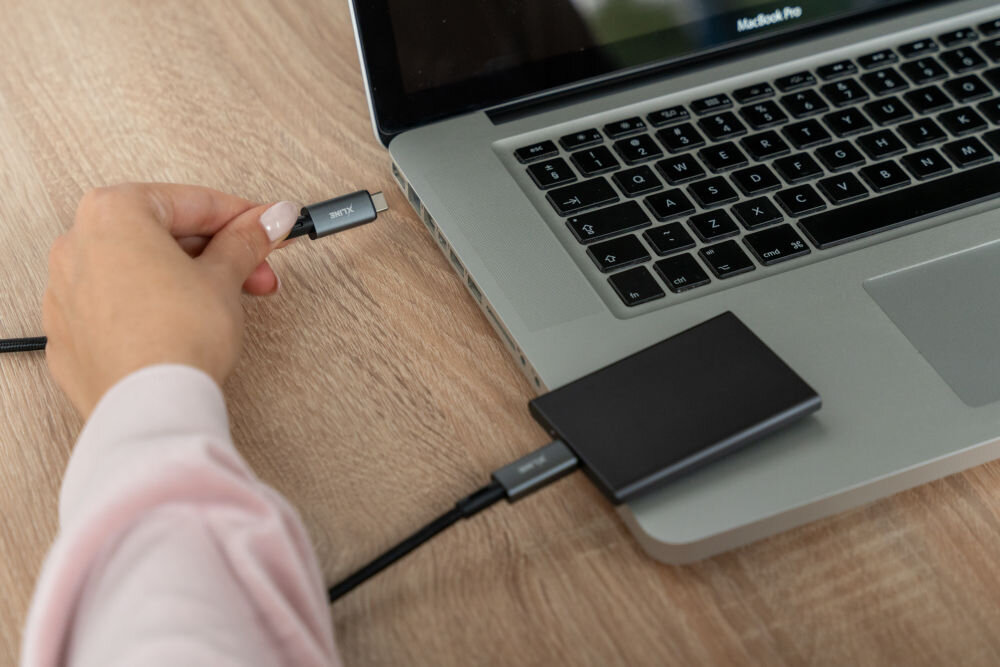 Osoba trzyma w ręku kabel USB-C, który jest podłączony do laptopa i zewnętrznego dysku twardego. Na ekranie laptopa widoczny jest napis \'MacBook Pro\'.