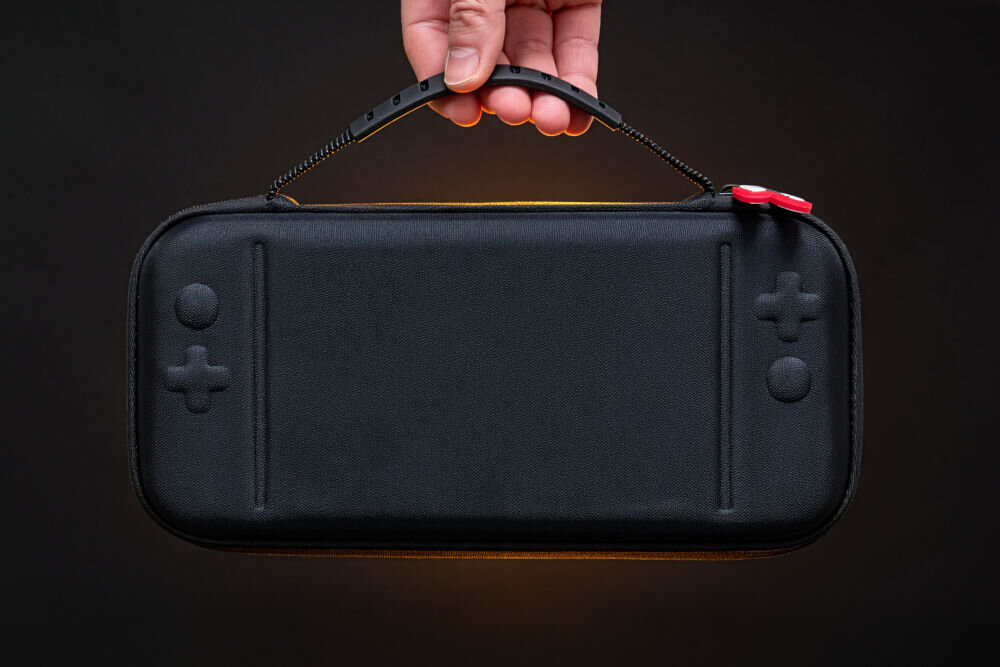 Etui MAD DOG NSC100K z wygodną rączką i paskiem do szybkiego wyciągania Nintendo Switch 2 Czarne, twarde etui na konsolę trzymane za czarny uchwyt na tle ciemnoszarym. Na powierzchni etui widoczne są tłoczenia imitujące kształt kontrolerów konsoli (krzyżak po lewej i przyciski po prawej). Widoczny również czerwono-czarny uchwyt zamka.