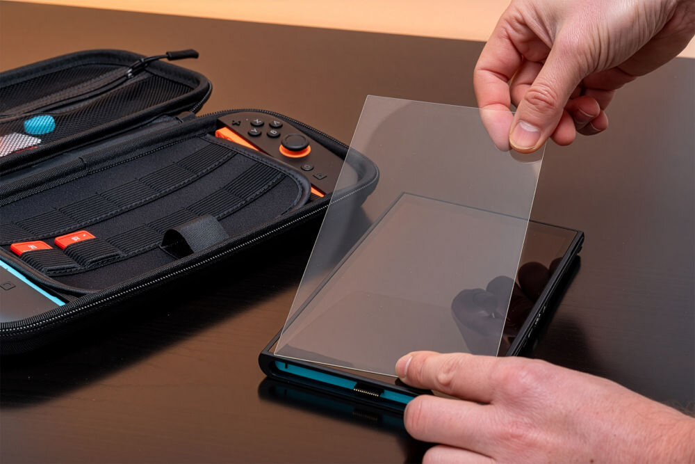 Etui MAD DOG NSC100K Dłonie nakładające przezroczystą folię ochronną na ekran czarnej konsoli leżącej w otwartym etui. W tle widoczne wnętrze etui z elastycznymi przegródkami na akcesoria i gry.