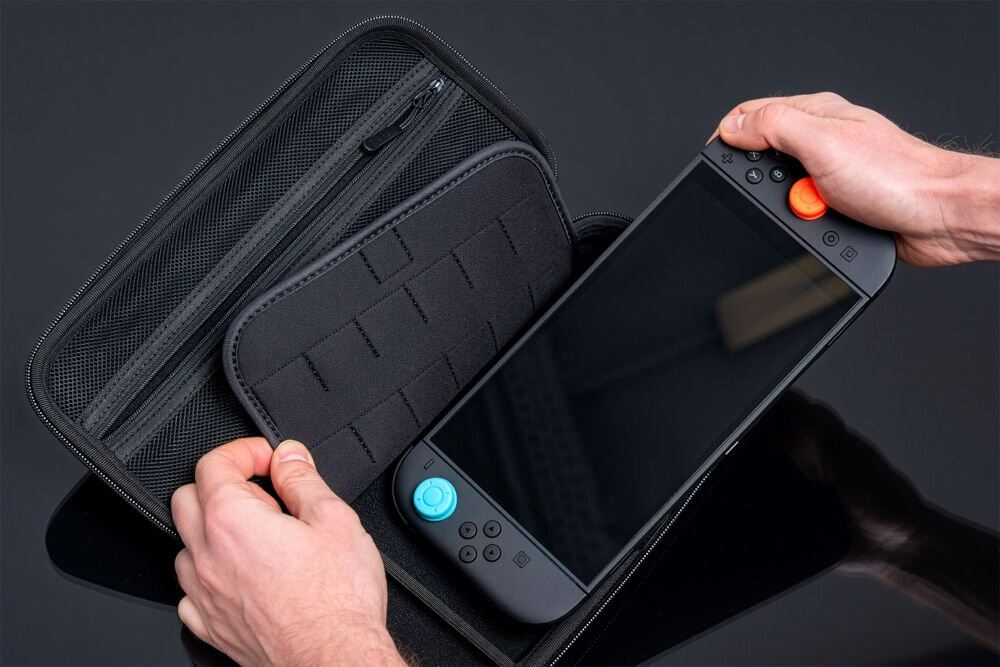 Etui MAD DOG NSC100K pancerny case dla Nintendo Switch 2 z wewnętrzną wyściółką Dłonie wyjmujące lub wkładające czarną konsolę (z niebieskim i pomarańczowym analogiem) do otwartego etui. W lewej części etui widoczna jest siatkowa kieszeń na zamek oraz gruba, czarna klapka z elastycznymi paskami do przechowywania gier.