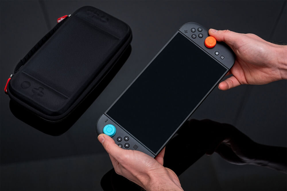 Etui MAD DOG NSC100K z miękką wyściółką chroniącą Nintendo Switch 2 przed zarysowaniami Dwie dłonie trzymają czarną konsolę, najprawdopodobniej Nintendo Switch, z wymienionymi na niebieski i pomarańczowy uchwytami analogów. Obok, na błyszczącej, czarnej powierzchni, leży zamknięte, czarne etui.