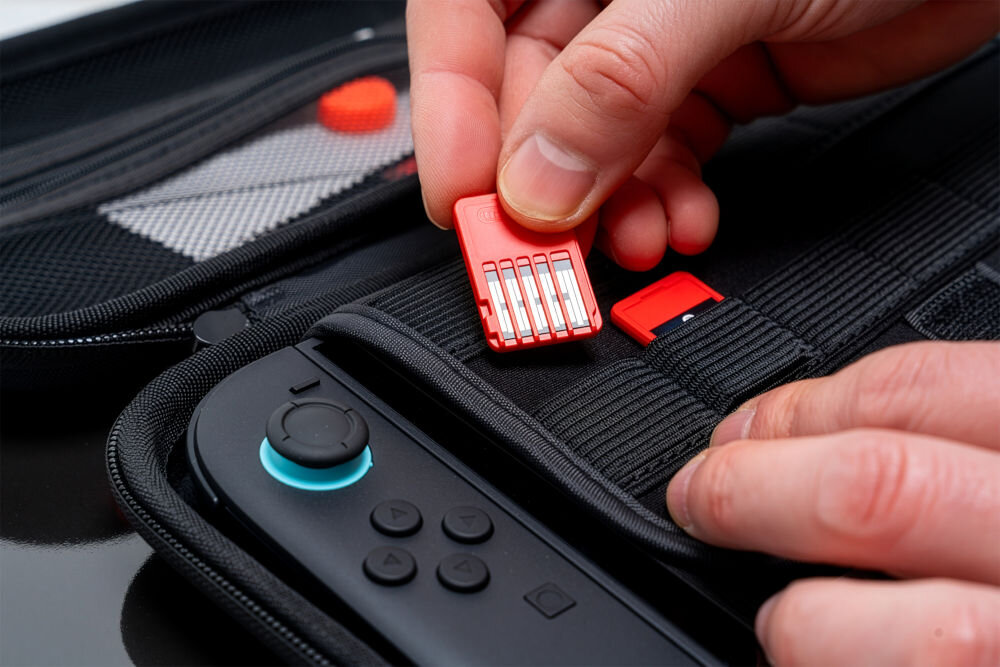 Etui MAD DOG NSC100K z wewnętrzną przegrodą na ładowarkę i karty pamięci dla Nintendo Switch 2 Zbliżenie na dłoń wkładającą czerwony kartridż z grą do jednej z elastycznych przegródek na gry wewnątrz otwartego, czarnego etui. W dole widoczna część konsoli z czarnym kontrolerem i niebieskim joystickiem.