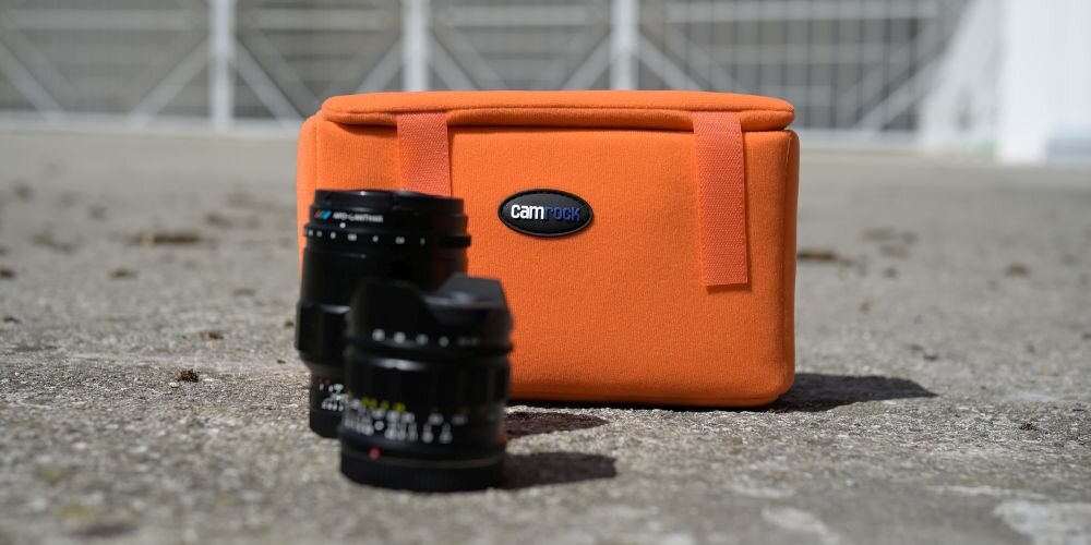 Na betonowej powierzchni znajduje się pomarańczowy wkład fotograficzny z logo \'camrock\', obok którego leżą dwa obiektywy fotograficzne.