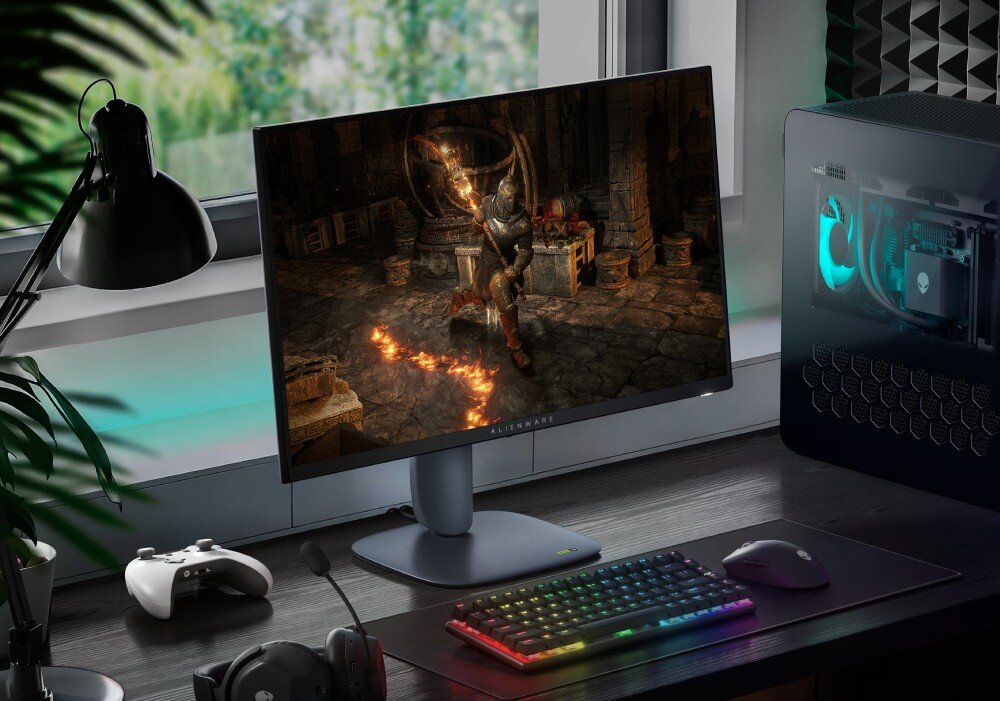 Monitor DELL Alienware AW2725DM Monitor wyświetlający grę fantasy stoi na biurku obok klawiatury z podświetleniem, myszki, słuchawek, kontrolera i komputera stacjonarnego