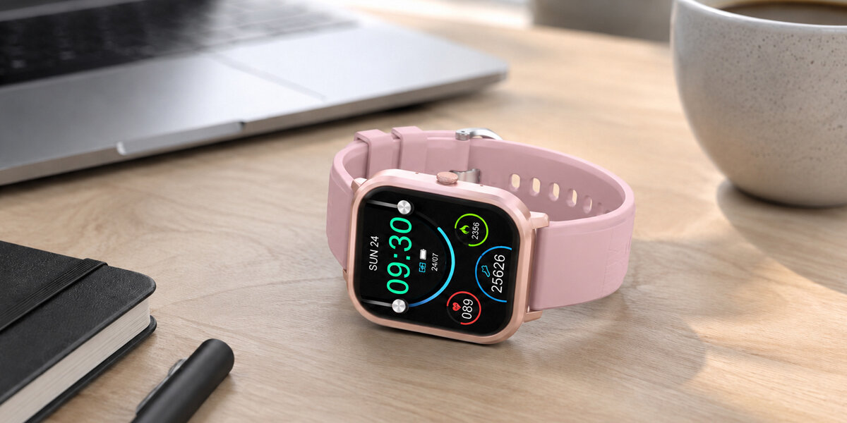 Różowy smartwatch Denver leżący na stole, obok widać laptop,notes,długopis oraz kubek z kawą.