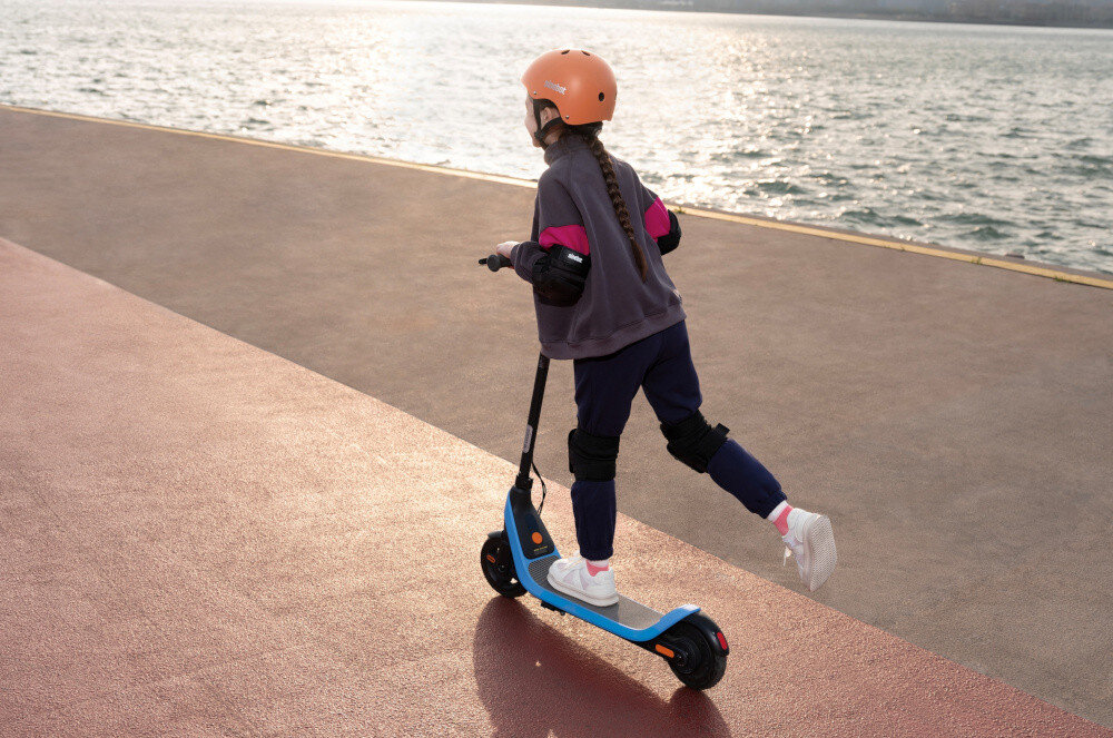 Hulajnoga elektryczna SEGWAY Ninebot C2 Lite 9km 130W 7 cali Czarno-niebieski chodnik droga dziecko jedzie na hulajnodze w tle woda rzeka morze wskaźnik LED można odczytać stan baterii i kontrolować poziom energii kierownica o wysokości 95 cm wygoda ergonomiczna pozycja antypoślizgowe uchwyty kontrola nad pojazdem bezpieczeństwo